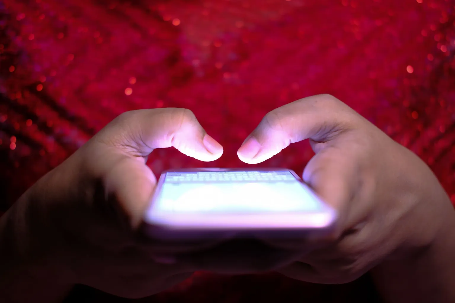Deux mains tenant un smartphone à l'écran lumineux, sur un fond rouge scintillant